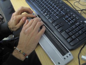 Close up of someone's hands as they read a braille display, with a keyboard nearby.